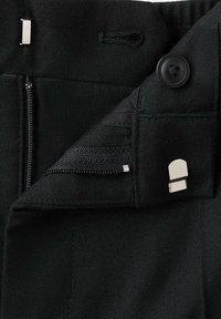 Gros plan de la ceinture d’un vêtement noir avec fermeture éclair, bouton et attaches métalliques à crochet et barre partiellement ouvertes.