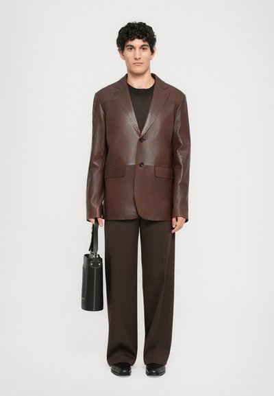 Man wearing dark brown leather blazer, matching pants, black shoes, and holding a black cylindrical handbag against white background.