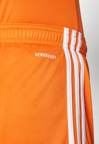 Pantalones cortos deportivos naranjas con marca AEROREADY, cinturilla elástica y tres rayas verticales blancas en el lateral. Textura de tela suave.