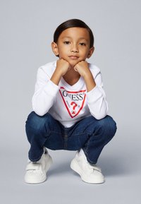 Camicia bianca a maniche lunghe con logo GUESS rosso, jeans in denim blu e sneakers bianche. Bambino accovacciato con le mani sul mento, sfondo neutro.
