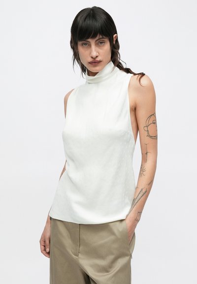 Valge varrukateta top kõrge kaelusega, millel on peen tekstuurimuster, kombineeritud beežide pükstega.