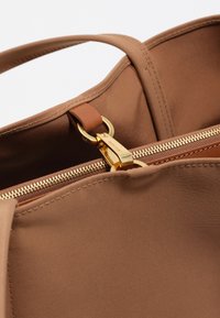 Brązowa torebka materiałowa wewnątrz z złotym suwakiem i skórzanym akcentem na pasku. Charakteryzuje się starannym ściegem i gładką teksturą.