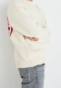 Suéter color crema en un punto texturizado, con un patrón geométrico rojo en el lateral. Combinado con jeans de mezclilla gris.