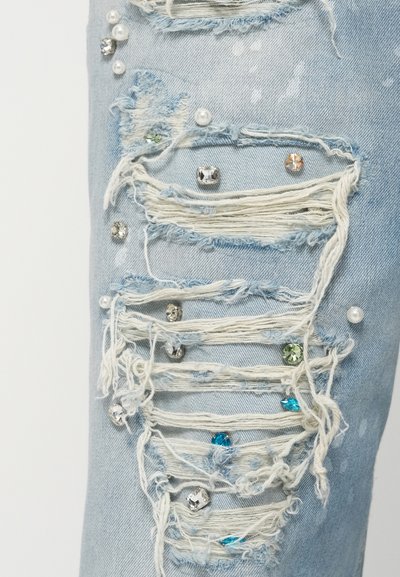 Poškodovane svetlo modre kavbojke z raztrganimi izrezi, okrašenimi z različnimi dragulji, vključno s srebrnimi, zelenimi in modrimi poudarki.