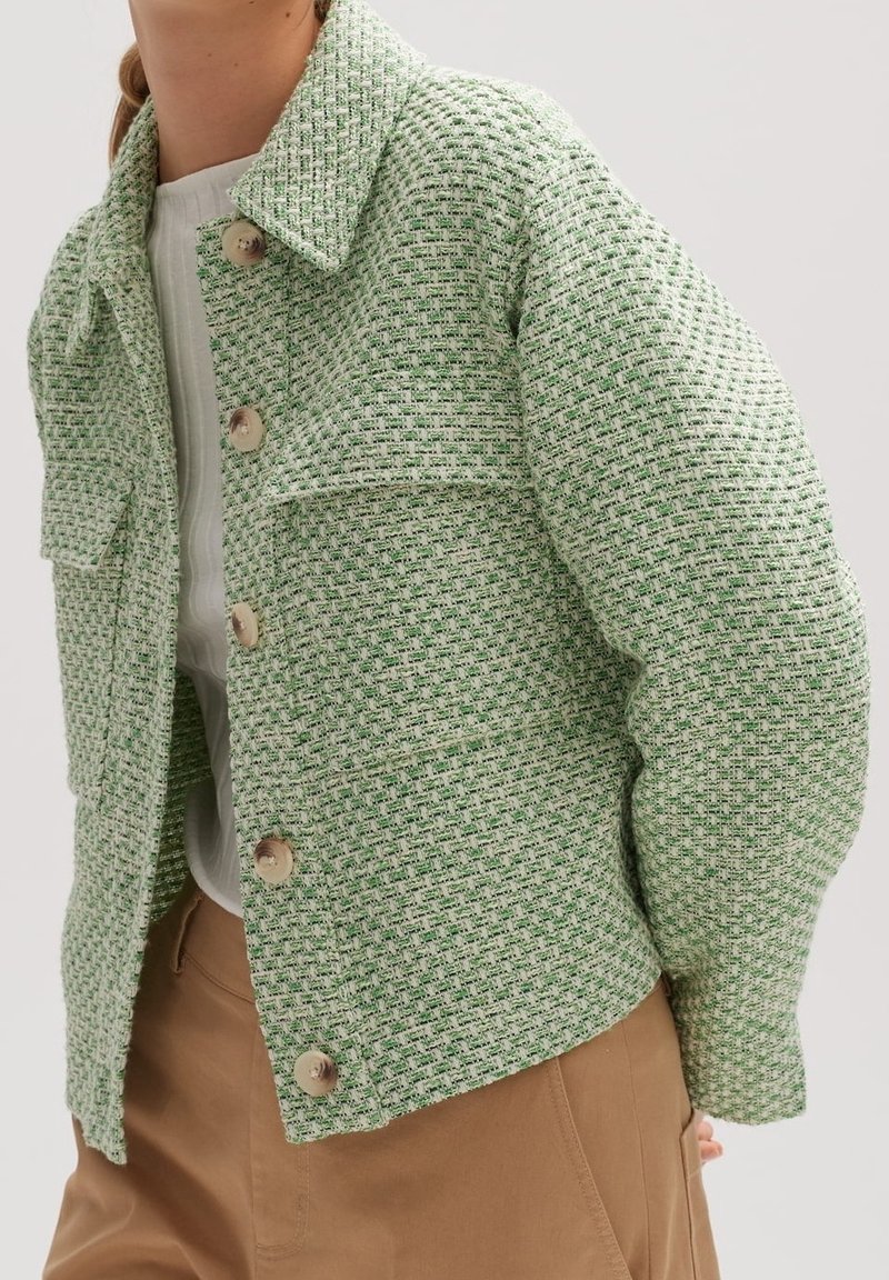 Grön texturerad jacka med ett vävt mönster, utrustad med krage, framfickor och stora knappar, kombinerad med beiga byxor.