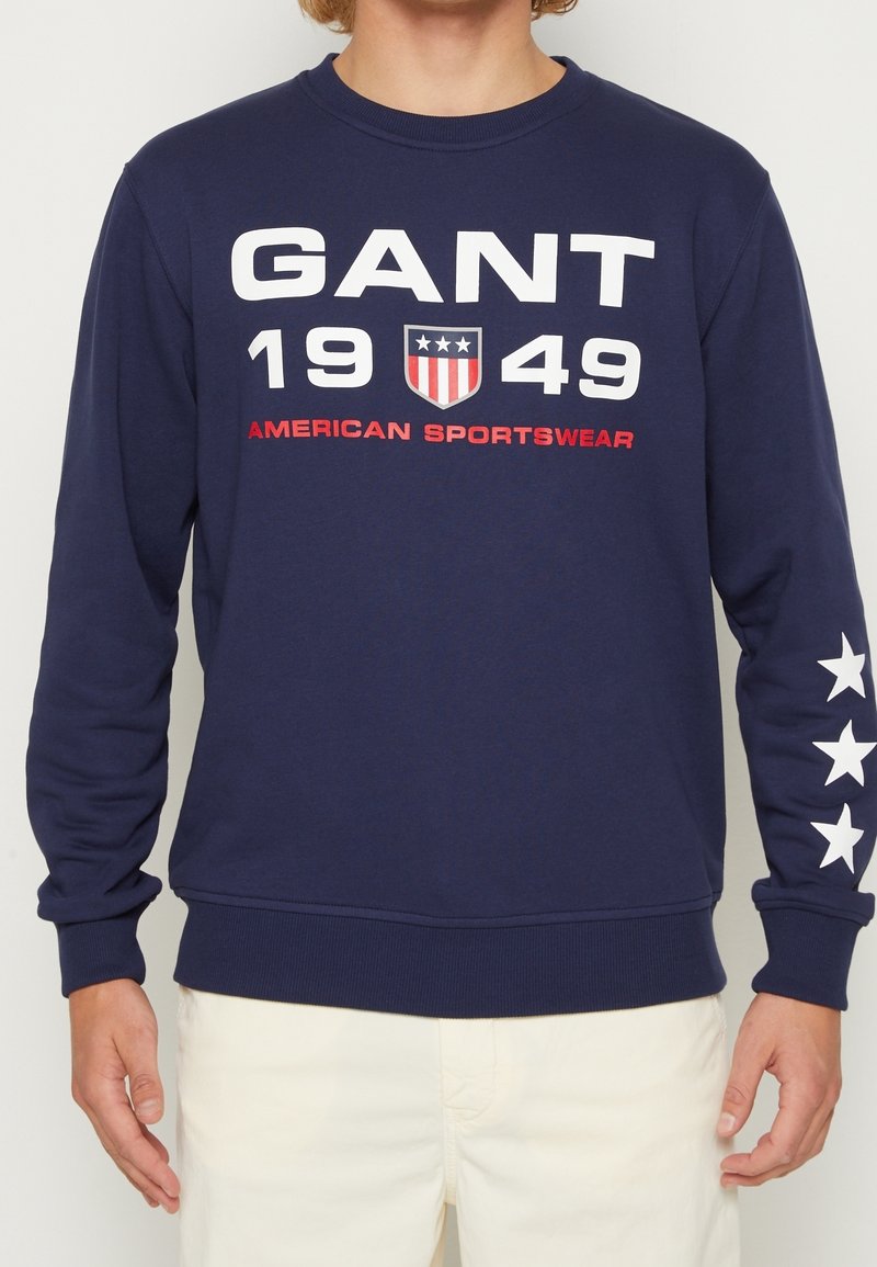 Marineblauwe sweatshirt van zachte stof met opvallende witte tekst "GANT 1949 AMERICAN SPORTSWEAR" en witte sterrenaccenten op de mouwen.