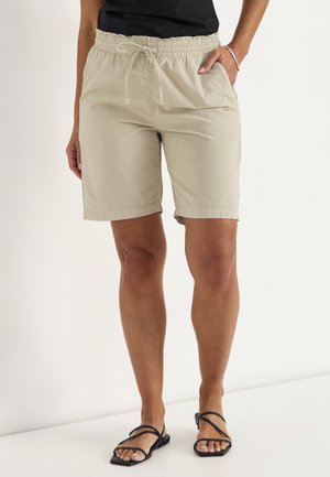 Vrouw draagt beige shorts met trekkoord, zwart mouwloos topje en zwarte sandalen met bandjes, staand met één hand in de zak.