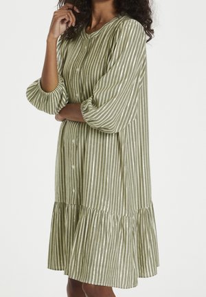 Woman wearing olive green and white vertical striped dress with long puffed sleeves and buttons down the front.