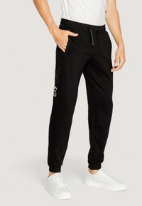 Black jogger pants with side pockets, drawstring waistband, and EA7 branding on the side; worn with white sneakers. Smooth fabric texture.
