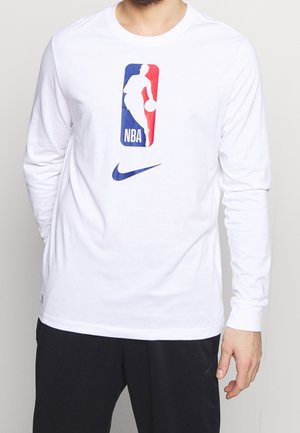 Person trägt ein weißes Langarmshirt mit NBA-Logo und blauem Nike-Swoosh, kombiniert mit schwarzer Hose, vor einfachem Hintergrund stehend.