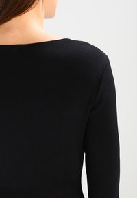 Haut noir à manches longues côtelé avec un large décolleté. Le matériau semble lisse, la coupe ajustée met en valeur la silhouette. Aucun motif ni accessoire visible.