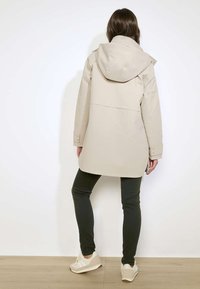 Vrouw die een beige cappuccino jas, zwarte broek en witte sneakers draagt, met haar rug naar de camera toe staand tegen een effen witte muur.