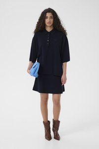 Polo supradimensionat bleumarin cu cinci nasturi, asortat cu o fustă în aceeași culoare. Geantă albastră texturată și ghete de gleznă din piele maro.