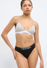 Γκρί μπραλέτα με λεπτές τιράντες και λευκή λωρίδα με λογότυπο, συνδυασμένη με μαύρο στρινγκ εσώρουχο με λευκή λωρίδα με λογότυπο, παρουσιάζοντας έναν απλό σχεδιασμό.