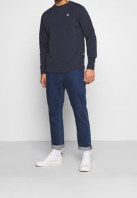 Marinblå långärmad sweatshirt med en diskret logotyp på bröstet, kombinerad med uppvikta mörkblå jeans och vita sneakers.