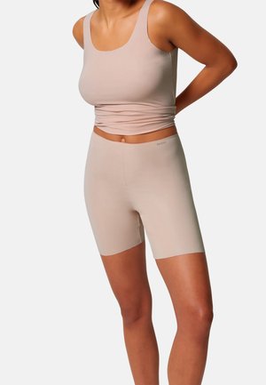 Frau trägt beige nahtloses Tanktop und hoch taillierte Biker-Shorts, steht mit den Händen hinter dem Rücken vor weißem Hintergrund.