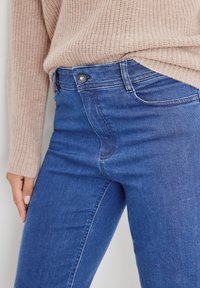 Jeans de mezclilla azul con un diseño ajustado, que cuentan con un cierre delantero de botones, cinco bolsillos y detalles de costura sutiles, llevados con un suéter de punto beige.