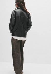 Schwarze Kunstlederjacke mit lässiger Passform, sichtbaren Nähten und Kragen, kombiniert mit lockeren braunen Hosen über einem weißen Hemd.