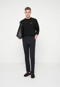 Maglietta nera a maniche lunghe con piccolo logo, pantaloni blu navy e mocassini marroni scuri. Tiene un grande borsa in pelle nera. Sfondo semplice.