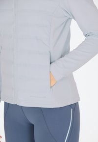 Lys grå isoleret jakke med et elegant design, der har lynlås foran og sidelommer, parret med mørkeblå leggings og kontrasterende syninger.