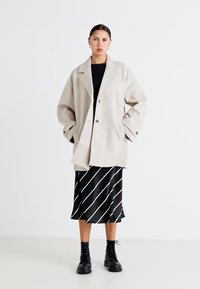 Beige ullblandede oversized kåpe med revers, to knapper og sidelommer, paret med et svart og hvitt stripete silkeskjørt og svarte støvler.