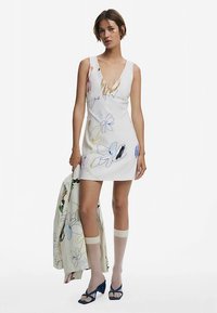 Witte mini-jurk met bloemenprint en diepe V-hals, gladde textuur, gecombineerd met een jas met bloemenprint en blauwe sandaaltjes met hak.