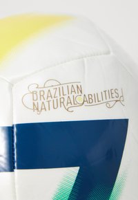 Biała piłka nożna z błyszczącym wykończeniem, z żółtymi gradientowymi akcentami, niebieskimi paskami i wytłoczonym napisem "BRAZYLSKIE NATURALNE ZDOLNOŚCI".