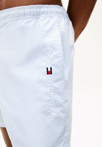 Shorts de bain bleu clair avec des rayures blanches et une taille élastique. Présente un logo blanc et des accents brodés rouge, blanc et bleu.