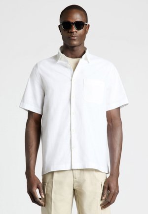 Uomo con occhiali da sole neri, camicia bianca a maniche corte con bottoni e pantaloni beige, in piedi davanti a uno sfondo chiaro e uniforme.