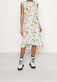 Blumenkleid in Hellblau mit Rüschenärmeln, tailliertem Bund und asymmetrischem Saum. Trägt dazu weiße Schnürstiefel mit schwarzen Sohlen.