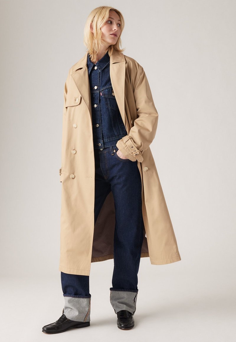 Trench coat beige con bottoni doppi, indossato sopra un giubbotto di jeans e jeans blu scuro, con maniche arrotolate e scarpe nere.