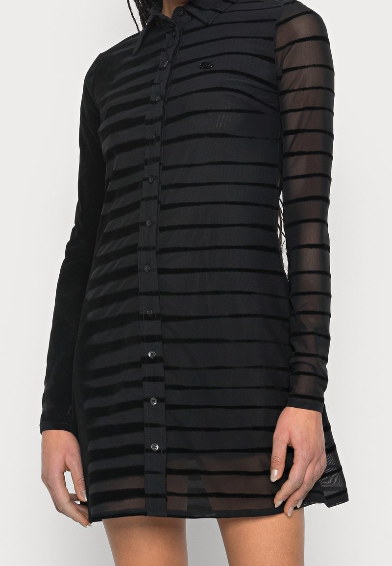 Femme portant une mini-robe noire transparente avec des bandes horizontales en velours, une fermeture à boutons sur le devant et des manches longues semi-transparentes.