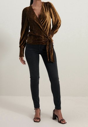 Blouse en velours froissé couleur bronze avec manches longues et ceinture à nouer à la taille, associée à un jean skinny noir et des talons sombres à brides.