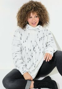 Weißer Strickpulli mit schwarzem Sprenkelmuster, hohem Kragen und langen Ärmeln. Kombiniert mit dunklen Jeans und schwarzen Stiefeletten.