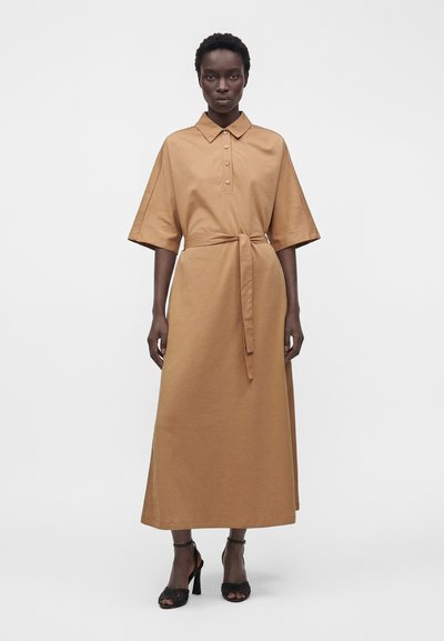 Femme debout portant une robe midi ample couleur beige avec manches courtes, col boutonné, ceinture en tissu et sandales à talons noires ouvertes à l'avant sur fond blanc.