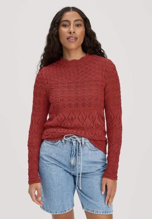 Femme portant un pull en maille texturée rouge à manches longues et un short en denim bleu clair taille haute avec une taille à cordon, debout devant un fond blanc.