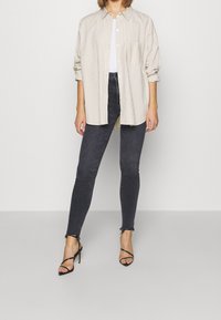 Striped beige oversized shirt with button closure, rolled sleeves, and a pocket. Paired with dark skinny jeans and black strappy heels.