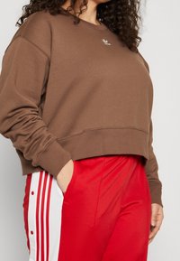 Kvinna med lockigt hår som bär en brun Adidas-pullover och röda byxor med vita sidostråk och knappar, handen i fickan.