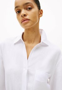 Camisa blanca de lino con cuello abotonado, un bolsillo en el pecho y tela texturizada, que presenta un ajuste relajado y detalles sutiles en las costuras.