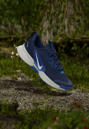 Zapatilla de trail running Nike azul y blanca en el aire sobre un camino de grava en un entorno boscoso, mostrando vistas lateral y inferior.