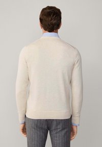 Suéter de punto de color beige claro con mangas largas, dobladillo y puños acanalados, combinado con pantalones gris de rayas, mostrando una camisa azul con cuello debajo.