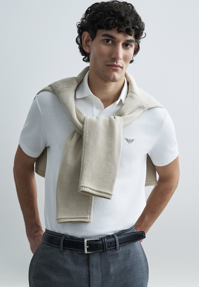 Pánsky outfit pozostávajúci z bielej polokošele, béžového pleteného svetra prehodeného cez ramená, sivých nohavíc a čierneho opasku so striebornou prackou.