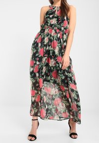Kvinna i ärmlös svart maxiklänning med blommor i rosa och vitt, kombinerad med svarta högklackade sandaler med ankelsnörning.