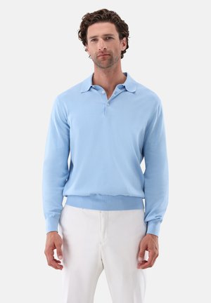 Mann mit kurzen lockigen braunen Haaren, der ein hellblaues Langarm-Poloshirt und weiße Hose trägt, steht vor einem schlichten Hintergrund.
