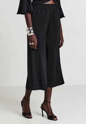 Donna che indossa ampi culotte neri, tacchi alti aperti neri, bracciali chunky argentati e anelli su uno sfondo neutro.