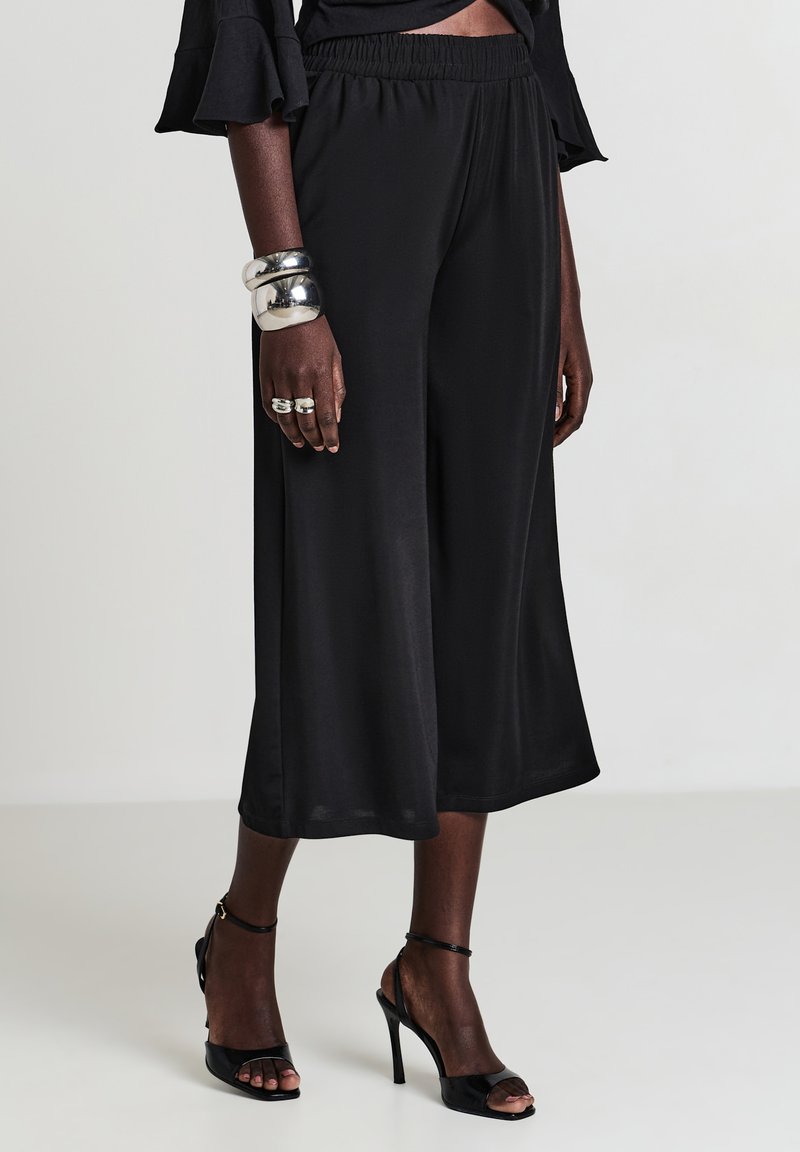 Frau in lockeren schwarzen Culottes, schwarzen offenen High Heels, silbernen klobigen Armbändern und Ringen vor einem schlichten Hintergrund.