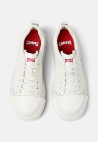White sneakers with a rounded toe, featuring lace-up design and a red interior. The brand name "CAMPER" is visible on the tongue.