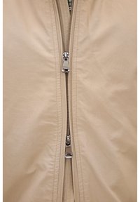 Hellbeige Jacke mit Reißverschluss und glatter Textur, mit einem zentralen Reißverschluss und einem Innenfutter, das am Kragen sichtbar ist.