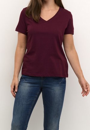 Vrouw draagt een bordeauxrode V-hals T-shirt met korte mouwen en een blauwe spijkerbroek, staand tegen een effen lichte achtergrond.