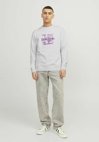 Grå sweatshirt med et lilla og sort grafisk logo på forsiden, parret med lyse, falmede jeans og hvide sneakers.